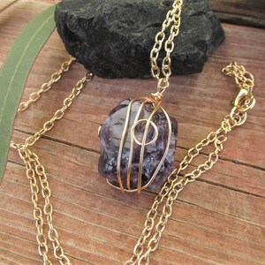 Puede incluir: Un collar de cadena dorada con una piedra de amatista p&uacute;rpura envuelta en alambre dorado. La piedra tiene una forma natural e irregular.
