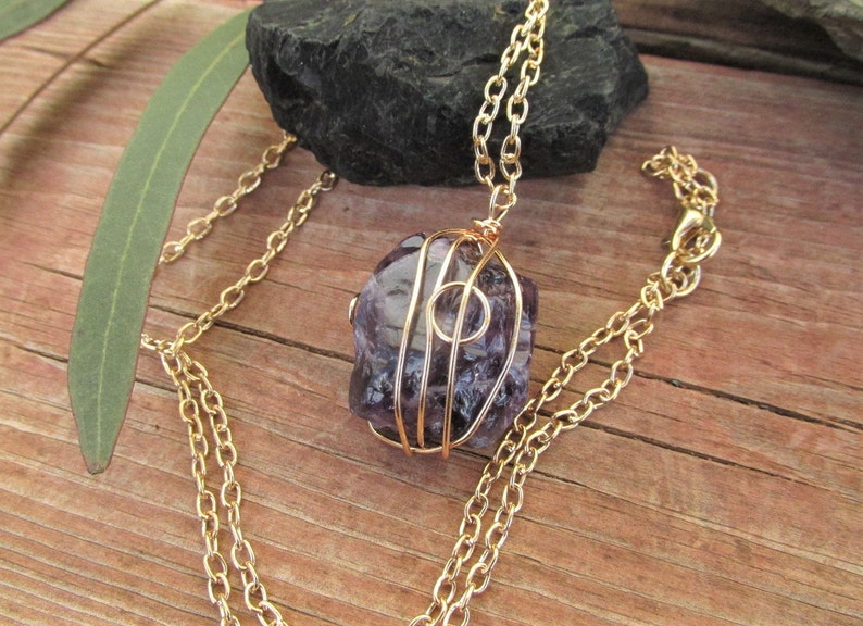 Puede incluir: Un collar de cadena dorada con una piedra de amatista p&uacute;rpura envuelta en alambre dorado. La piedra tiene una forma natural e irregular.