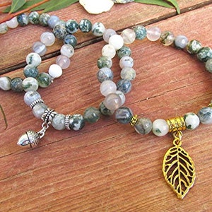 May include: Two green and white agate stone bracelets with silver acorn and gold leaf charms. The bracelets are on a wooden surface.