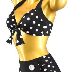 May include: Black and white polka dot bikini with a high-waisted bottom featuring white buttons. The top has a bow tie detail. The bikini is on a gold mannequin.