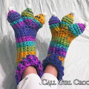 May include: A pair of colorful, crocheted claw slippers. The slippers feature a rainbow of colors, including purple, blue, green, and yellow. Each claw has a white pom-pom at the tip. The text "Call Girl Crochet" is visible.