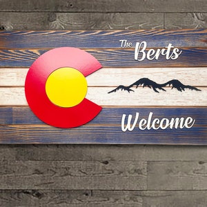 Puede incluir: Un letrero de madera con un acabado de pintura azul y blanca desgastada. El letrero presenta un diseño de bandera de Colorado rojo, amarillo y blanco, una silueta de montañas y el texto "The Berts Welcome".