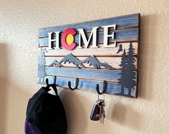 May include: A rectangular wooden wall-mounted key holder with a distressed blue and white finish. It features the word HOME with a Colorado state flag emblem as the letter O, a black mountain silhouette, pine trees, and three metal hooks.
