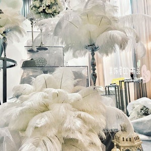 May include: White ostrich feathers arranged in a vase on a table with white balloons and a floral centerpiece. The feathers are tall and fluffy, creating a dramatic and elegant look.