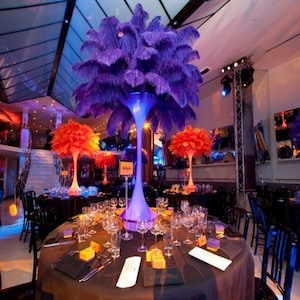 May include: A table setting with black tablecloths and purple and orange feather centerpieces. The centerpieces are tall and have a modern design. The table is set with glasses, silverware, and place cards.