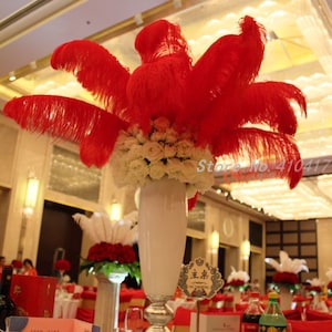 Puede incluir: Un centro de mesa de boda con un jarrón blanco lleno de rosas blancas y plumas de avestruz rojas. La mesa está puesta con manteles rojos, vasos y una botella de refresco.
