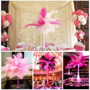 May include: Pink and white ostrich feathers arranged in a vase as a centerpiece for a wedding or party. The feathers are displayed on a table with pink and white tablecloths and chairs. The background is a white sheer curtain with twinkling lights.