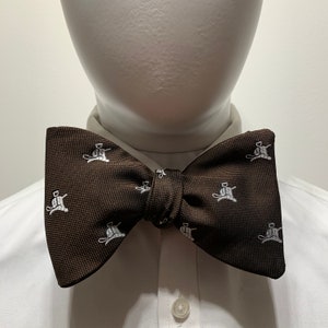 Könnte beinhalten: Eine braune Fliege mit einem weißen Muster aus Strichmännchen, die Hockey spielen. Die Fliege ist gebunden und wird auf einem weißen Hemd mit Kragen getragen.