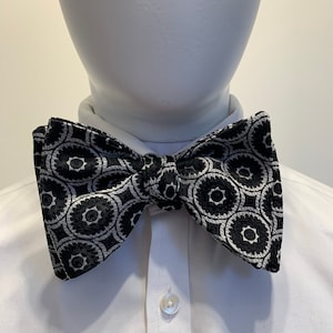 Könnte beinhalten: Eine schwarz-weiße Fliege mit geometrischem Muster. Die Fliege ist aus einem strukturierten Stoff gefertigt und hat ein kreisförmiges Design.