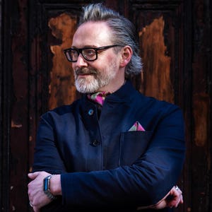 May include: A man wearing a navy blue jacket with a pink and green patterned pocket square and scarf. He is wearing black-framed glasses and a silver watch. The background is a dark wooden door.