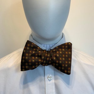 Könnte beinhalten: Eine braune Fliege mit einem Muster aus kleinen gelben und grünen Kreisen. Die Fliege ist um einen weißen Hemdkragen gebunden.