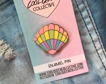 May include: A gold enamel pin featuring a rainbow seashell with a pink, yellow, green, and purple colour scheme. The pin is on a white card with the text "Cool Queen Collective" and "Enamel Pin" printed on it.
