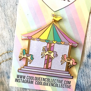 May include: A colorful carousel pin with a gold outline. The carousel has a rainbow-colored roof and three pastel-colored horses. The pin includes the text "WWW.COOLQUEENCOLLECTIVE.COM" and "INSTAGRAM: COOLQUEENCOLLECTIVE".