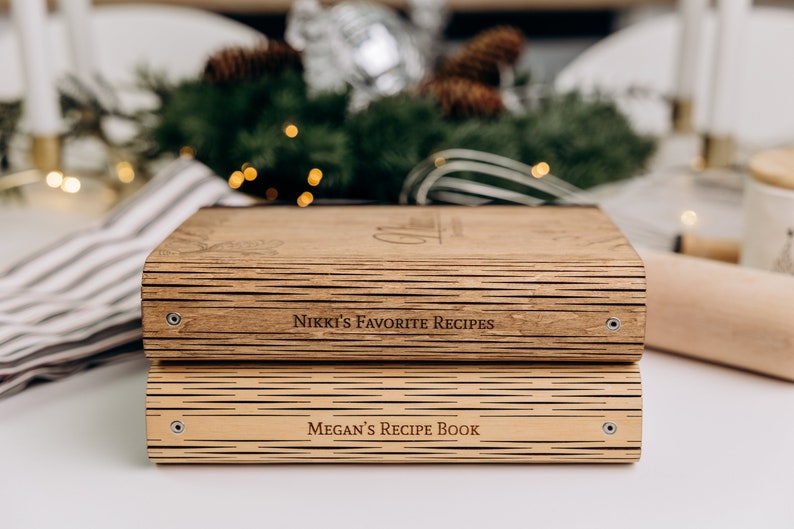 May include: Two wooden recipe books stacked on top of each other. The top book is labeled "Nikki's Favorite Recipes" and the bottom book is labeled "Megan's Recipe Book".