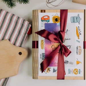 May include: A wooden box with a burgundy ribbon tied around it. The box is decorated with various kitchen-themed illustrations and the text "Let's roll", "My Kitchen", "Cook it Easy", "Baked", "Kitchen Queen", "Kitchen Blessed", and "Love".