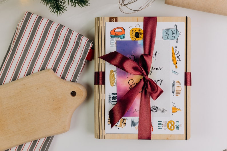 May include: A wooden box with a burgundy ribbon tied around it. The box is decorated with various kitchen-themed illustrations and the text "Let's roll", "My Kitchen", "Cook it Easy", "Baked", "Kitchen Queen", "Kitchen Blessed", and "Love".
