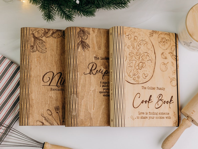 May include: Three wooden recipe books with a rustic design. The books are titled "The Collier Family Cookbook", "Our Favorite Recipes", and "Nick's Favorite Recipes". The covers feature a variety of illustrations, including a pizza, tomatoes, and kitchen utensils.