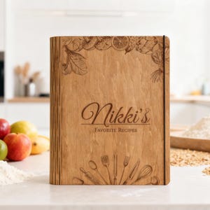 Peut inclure: Un classeur de recettes en bois avec le texte "Nikki's Favorite Recipes". La couverture présente des illustrations gravées de fruits, de légumes et d'ustensiles de cuisine. Le classeur est sur une surface blanche avec des fruits et de la farine.