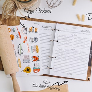 May include: A brown leather recipe binder with a white interior and lined pages. The binder has a ring mechanism and a tab labeled "Breakfast". The binder is surrounded by various kitchen items, including a rolling pin, pasta, and a wooden cutting board. The image also includes a sheet of stickers with various kitchen-themed designs.