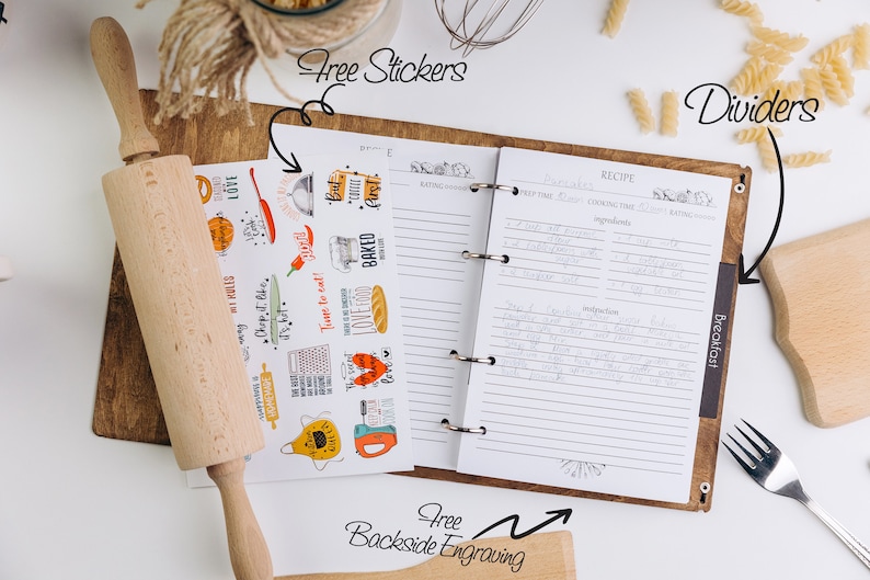 May include: A brown leather recipe binder with a white interior and lined pages. The binder has a ring mechanism and a tab labeled "Breakfast". The binder is surrounded by various kitchen items, including a rolling pin, pasta, and a wooden cutting board. The image also includes a sheet of stickers with various kitchen-themed designs.