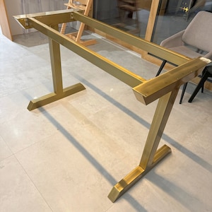 May include: A gold metal table base with a simple, modern design. The base is made of two rectangular pieces of metal that are joined together in a T-shape. The base is sturdy and well-made, and it would be a great addition to any dining room or kitchen.