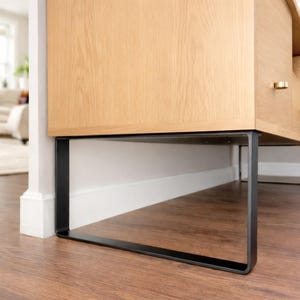 Op de afbeelding: Een lichtgekleurde houten kast met een zwart metalen frame. De kast heeft een rechthoekige vorm en staat tegen een witte muur en een houten vloer. Het ontwerp van de kast is modern en minimalistisch.