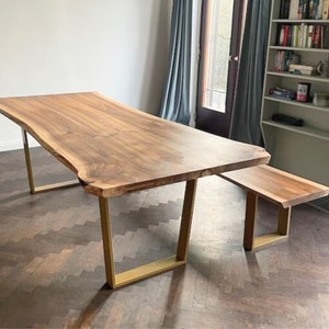 May include: A large wooden dining table with a live edge top and gold metal legs. The table is paired with a matching wooden bench with gold metal legs.