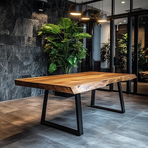 Op de afbeelding: Een grote houten tafel met een live edge blad en zwarte metalen poten. De tafel staat in een moderne woonkamer met een groene plant op de achtergrond.