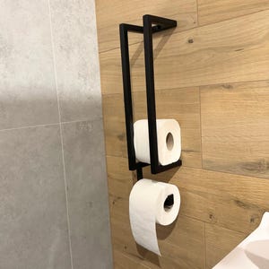 May include: A black metal toilet paper holder with two rolls of white toilet paper. The holder is mounted on a wooden wall. The design is modern and minimalist, suitable for a bathroom.