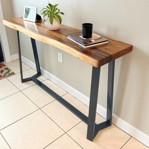 Pode incluir: Uma mesa de consola de madeira com tampo de borda viva e pernas de metal preto. A mesa é decorada com uma pequena planta em vaso, um porta-retratos e um livro.