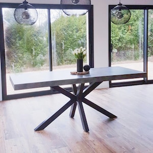 May include: A gray concrete dining table with a black metal X-shaped base. The table is set with a vase of white flowers and a wooden tray with a decorative object.