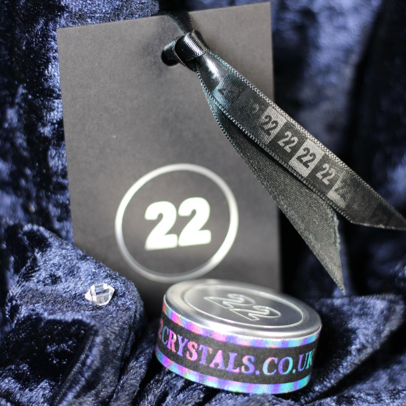 May include: A black tag with a white circle containing the number 22. A black ribbon with the number 22 printed repeatedly is tied around the tag. A silver container with a holographic sticker that reads "CRYSTALS.CO.UK" is in the foreground.