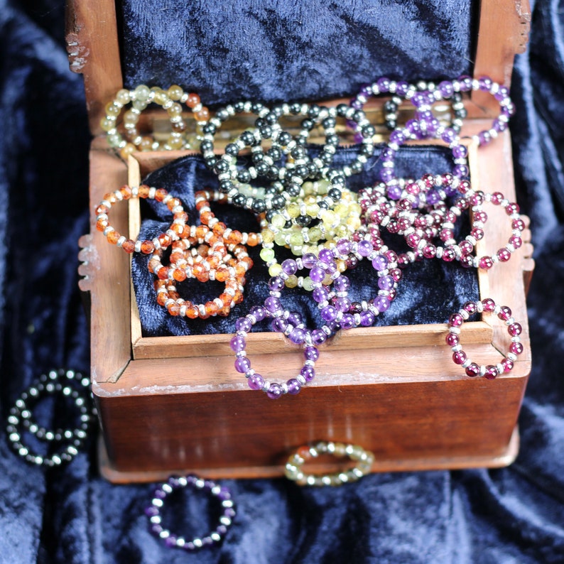 May include: A wooden box overflowing with colorful gemstone bracelets. The bracelets are made of various stones, including amethyst, carnelian, and citrine. The box is lined with a dark blue velvet fabric.