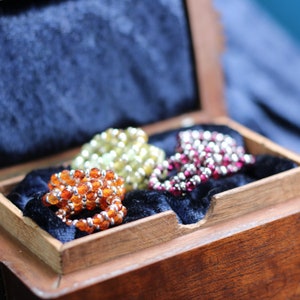 May include: Three beaded rings in a wooden box. The rings are made of different colored beads, including orange, yellow, and pink.