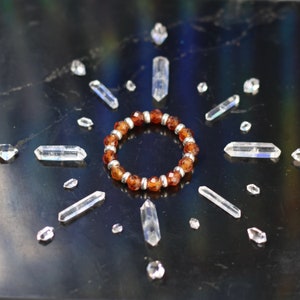 May include: A brown gemstone bracelet with silver beads, surrounded by clear quartz crystals on a black background.
