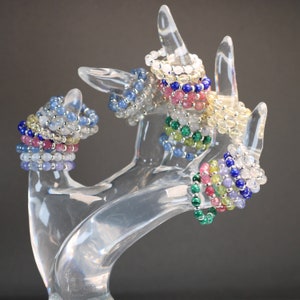 May include: A set of colorful beaded bracelets made with various gemstones, including blue, green, pink, and white. The bracelets are stacked on a clear plastic hand-shaped display stand.