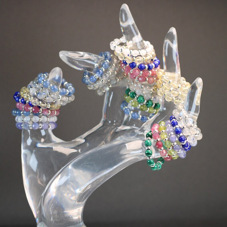 May include: A set of colorful beaded bracelets made with various gemstones, including blue, green, pink, and white. The bracelets are stacked on a clear plastic hand-shaped display stand.