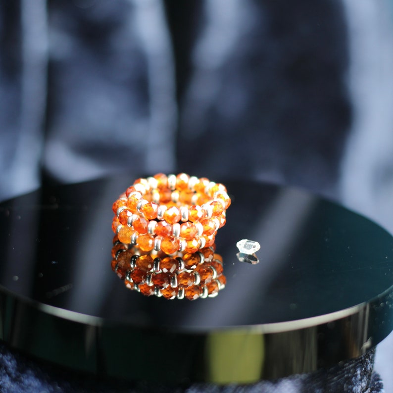 May include: A close-up of a stack of orange gemstone beads with silver accents on a black surface. The beads are arranged in a circle and reflect the light.