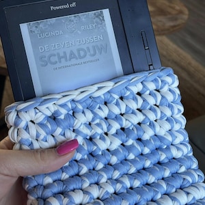 Puede incluir: Un lector electrónico en una funda de ganchillo azul y blanco. El lector muestra el texto "DE ZEVEN ZUSSEN SCHADUW" de Lucinda Riley. La funda es de tela gruesa y tejida.