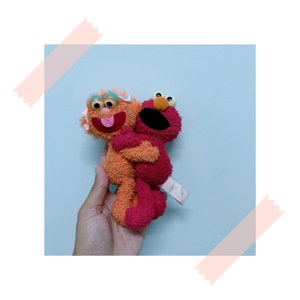 May include: Orange and red plush Sesame Street characters, Elmo and Abby Cadabby, hugging each other.