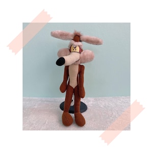 May include: A brown and tan plush toy of Wile E. Coyote from the Looney Tunes cartoon series. The coyote is wearing a red and white pilot's helmet and goggles. The toy is standing on a black stand.