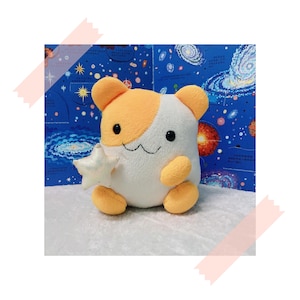 Può includere: Un giocattolo di peluche a forma di gatto con orecchie e zampe arancioni, pelliccia bianca e occhi neri. Il giocattolo tiene una piccola stella bianca nelle sue zampe.
