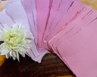 Puede incluir: Hojas de papel hechas a mano en tonos rosa y lavanda, con un borde rugoso y texturizado. Una gran flor de crisantemo blanca y amarilla se coloca cerca del papel, creando un contraste de texturas y colores.
