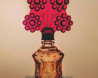 Puede incluir: Una botella de vidrio vintage con un tapón de vidrio rojo en forma de árbol estilizado con adornos redondos de vidrio rojo. La botella es transparente y tiene una superficie texturizada.