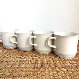 May include: Four white ceramic mugs with a light blue stripe around the top. The mugs are sitting on a woven wicker surface.