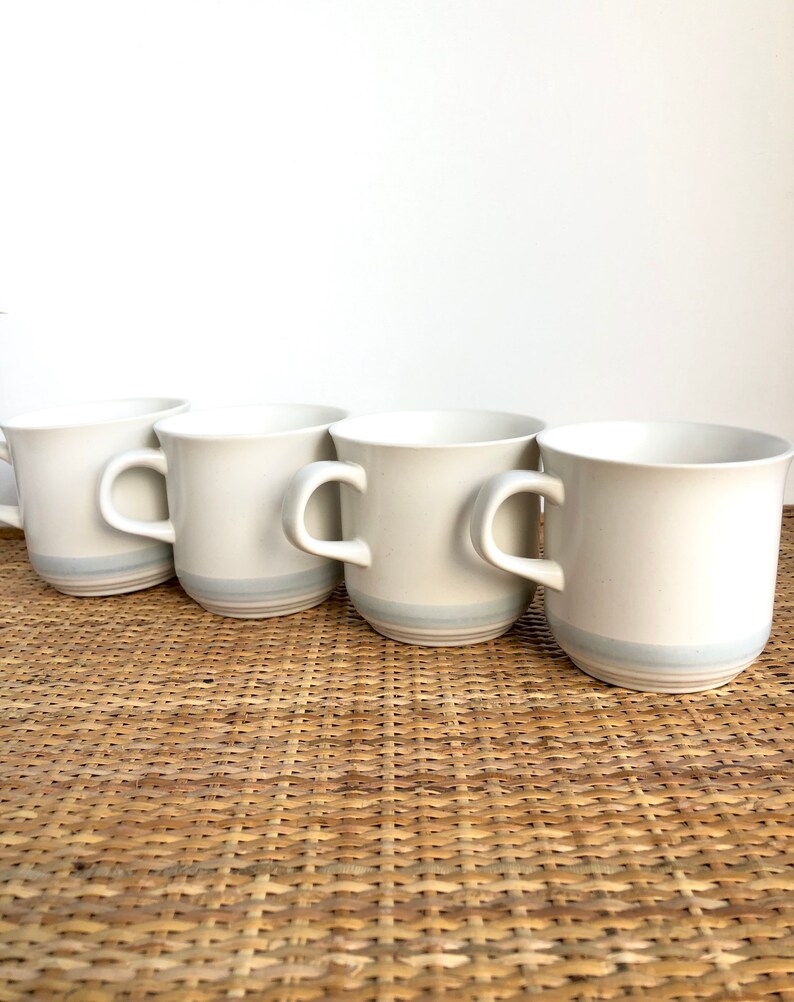 May include: Four white ceramic mugs with a light blue stripe around the top. The mugs are sitting on a woven wicker surface.
