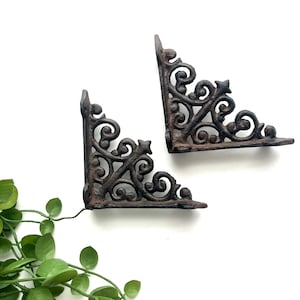 May include: Two dark brown cast iron shelf brackets featuring an intricate scrollwork pattern with curved lines and a pointed top detail. The brackets have a textured, weathered finish and are positioned at an angle against a plain background.