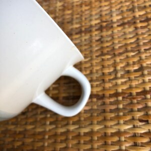 May include: A white ceramic teacup with a handle. The cup is sitting on a woven wicker surface.