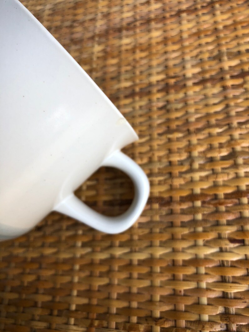 May include: A white ceramic teacup with a handle. The cup is sitting on a woven wicker surface.