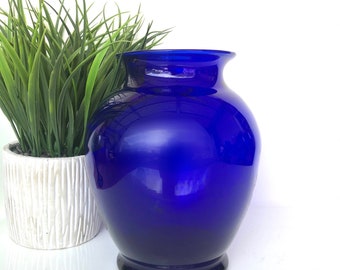 May include: A vibrant cobalt blue glass vase with a rounded body and a flared rim. The vase has a dark base and sits next to a white textured planter with green grass. The vase is a decorative home decor item.